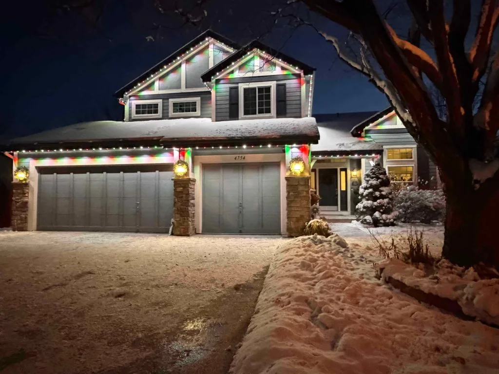 A snow-covered house adorned with colorful holiday lights glows softly at night