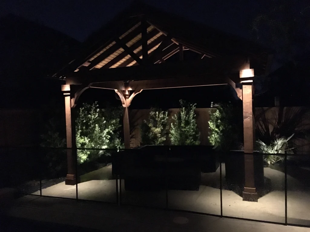A dimly lit wooden gazebo is surrounded by softly illuminated greenery at night